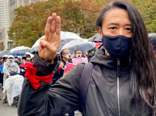 雨の中、都内でデモに参加する保芦ヒロスケさん。アウンサンスーチー氏が所属する国民民主連盟（NLD）の政党カラーである赤色のリボンを手首に巻く（保芦さん提供）